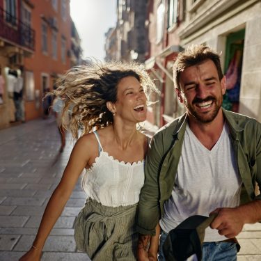 Young cheerful couple having fun while holding hands and running in spring day on the city street.